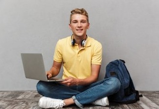 Portrait of a smiling casual teenage boy with backpack using laptop computer while sitting isolated over gray background