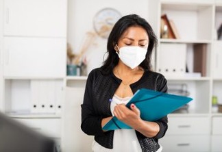 Latin american female business assistant wearing medical face mask standing in office with clipboard, noting tasks. Concept of social distance in coronavirus pandemic