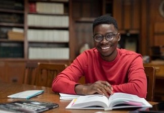 Portrait of university student doing homework in school library and smiling. Happy high school student looking at camera while studying for exam. African american clever guy with open book sitting at desk with copy space.