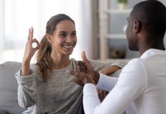 Happy mixed race deaf and dumb couple using sign language for communication at home sit on sofa, interracial hearing impaired disabled man and woman showing hand gestures as finger speech concept