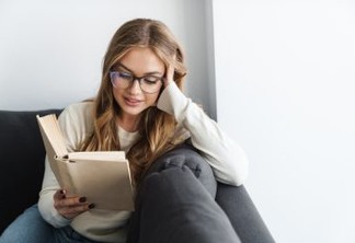 Photo of young pleased woman in eyeglasses reading book while sitting on couch at living room