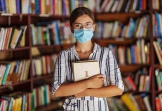 Portrait of young attractive college girl standing in library with face mask on holding a book. Studying during covid 19 concept.