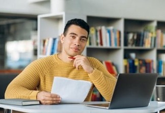Portrait of young adult hispanic male freelancer or student is looking at the camera with smile. Attractive business man sits at the work desk, making notes, learning online
