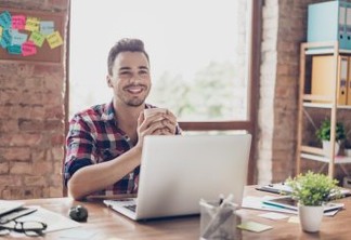 Low angle shot of successful happy brunet student, sitting with cup of tea, smiling, in front of computer, in casual checkered wear, at his work station in modern light office