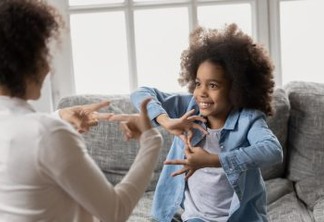 Smiling little african american girl with hearing loss practicing body language with professional female therapist at home, happy deaf kid communicating nonverbal with mum using signs and gestures.
