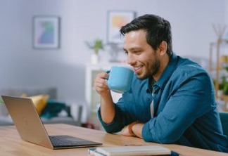 Portrait of Smiling Young Man Working on a Laptop from Home and Drinking Coffee From the Mug and is Satisfied with His Work. Cozy Modern Flat done in Scandinavian Style.
