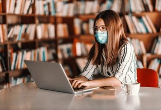 Young attractive female student with brown hair having face mask and using laptop while sitting in library. Remote learning concept.