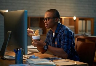 Profile view of handsome young man eating instant noodles with chopsticks while watching favorite TV series on computer, blurred background