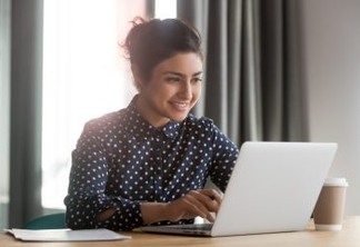 Happy young indian business woman entrepreneur using computer looking at screen working in internet sit at office desk, smiling hindu female professional employee typing email on laptop at workplace