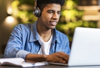 Smiling african american teen guy in headphones looking at laptop, studying foreign language through video conference application, cafe interior