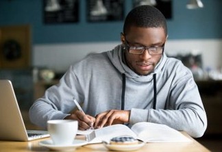 Focused millennial african american student in glasses making notes writing down information from book in cafe preparing for test or exam, young serious black man studying or working in coffee house