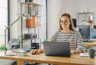 Beautiful Young Woman in Glasses is Working on a Laptop in a Creative Business Agency. They Work in Loft Office. Diverse People Working in the Background. She's in a Good Friendly Mood.
