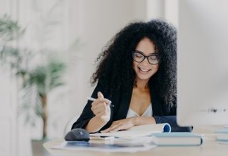 Photo of pleased curly haired woman writes down some information, holds pen, has smile, wears optical glasses and formal clothes poses at workplace with computer. Student writes ideas for course paper