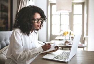 Young African American girl writing notes in restaurant.Nice girl with dark curly hair sitting in cafe with laptop and notebook.Portrait of lady in glasses thoughtfully looking in laptop with notebook