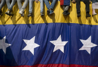 In this Oct. 26, 2016 photo, people sit on a wall draped by a Venezuelan flag during a protest against President Nicolas Maduro in Caracas, Venezuela. Venezuela's standoff deepened after congress voted to open a political trial against Maduro for breaking the constitutional order and opposition leaders called for mass demonstrations on Wednesday to drive the leader from office. (AP Photo/Alejandro Cegarra)