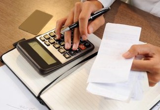 Closeup of person holding bills and calculating them. Notebook, calculator and credit card lying on desk. Payment concept. Cropped view.