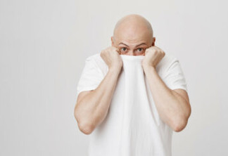 Indoor shot of funny bald caucasian man hiding head in his white t-shirt and glancing at camera while standing over gray background. Guy is embarrassed and trying to escape from people who look at him.